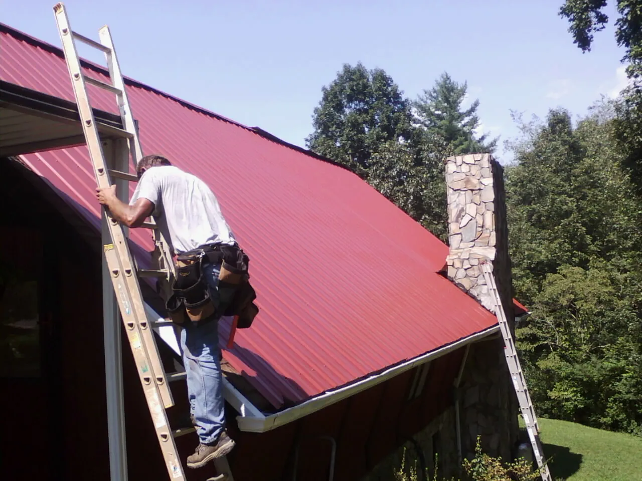 Roofer on a ladder beside a red metal roof and stone chimney — Soffit & Fascia Repair by Roofing Brothers in Merion Station